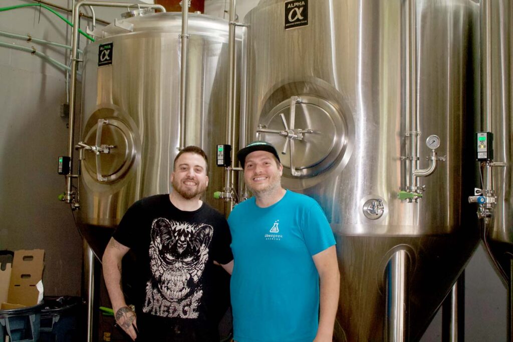 A man in a black tshirt stands arm in arm with another man in a blue tshirt. Both are smiling and standing in front of tall, silver beer brewing tanks.