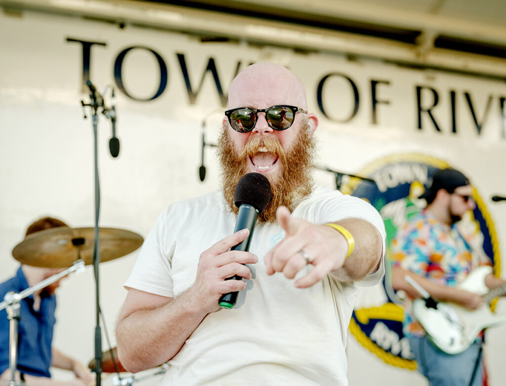 A man with a red beard and wearing sunglasses points to the camera while singing into a microphone during a concert on the Riverhead main stage of the Alive on 25 block party.