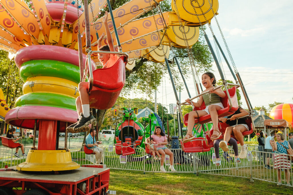 A brown-haired girl screams with joy on a carnival ride. The boy in front of her holds his hands in the air enjoying the swings ride.