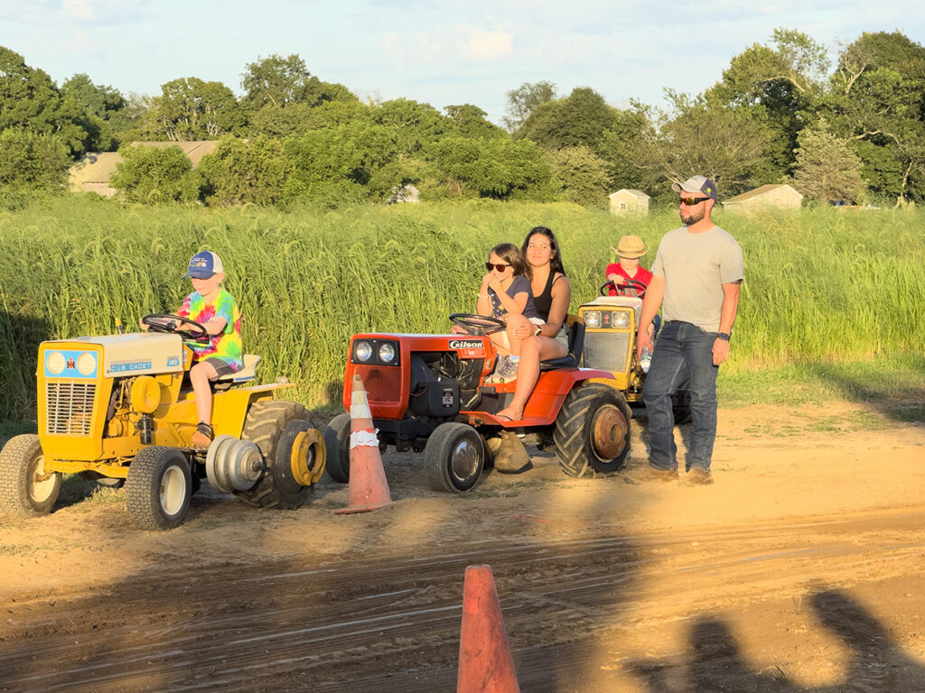 A woman in a black shirt holds a young girl on her lap while sitting on their red garden tractor, waiting for their turn to pull the weighted sled.