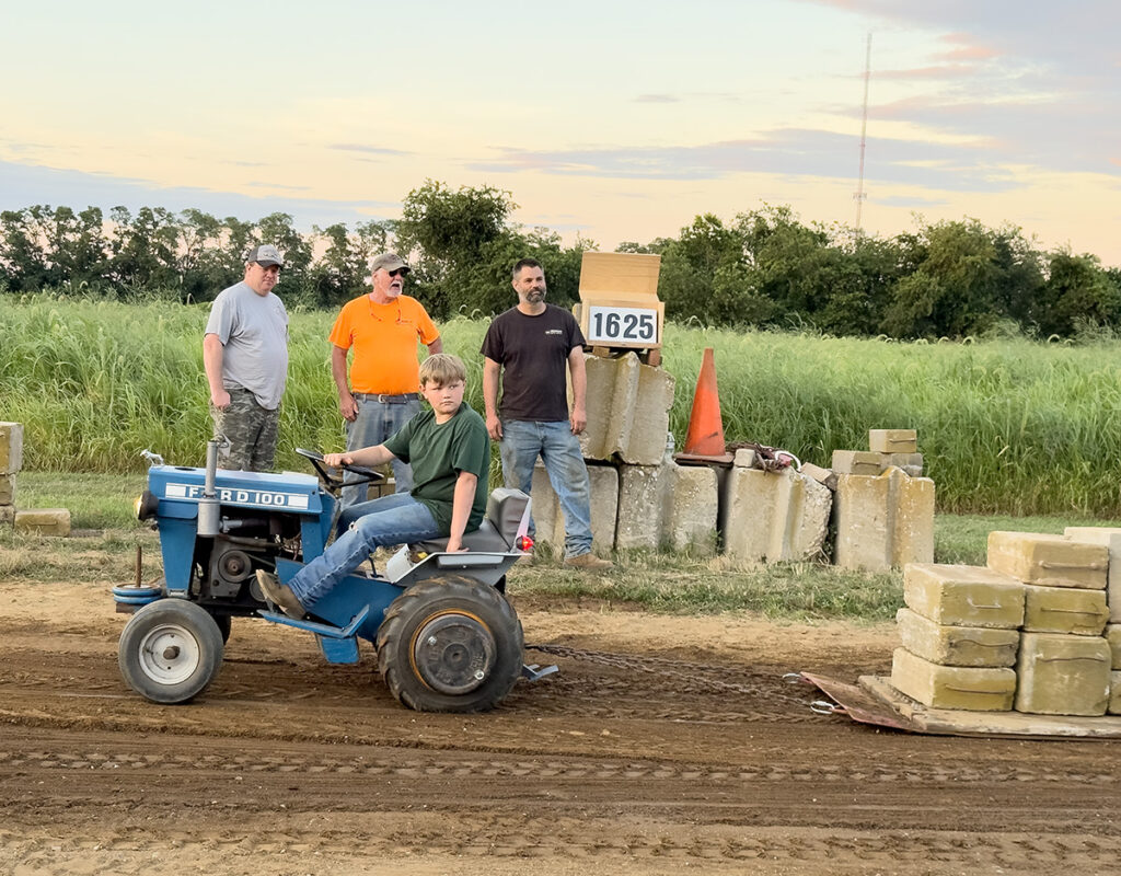A boy on a small blue tractor pulls 1625 pounds of concrete blocks on the sled behind his tractor. Three men stand in the background waiting to see if he makes it.