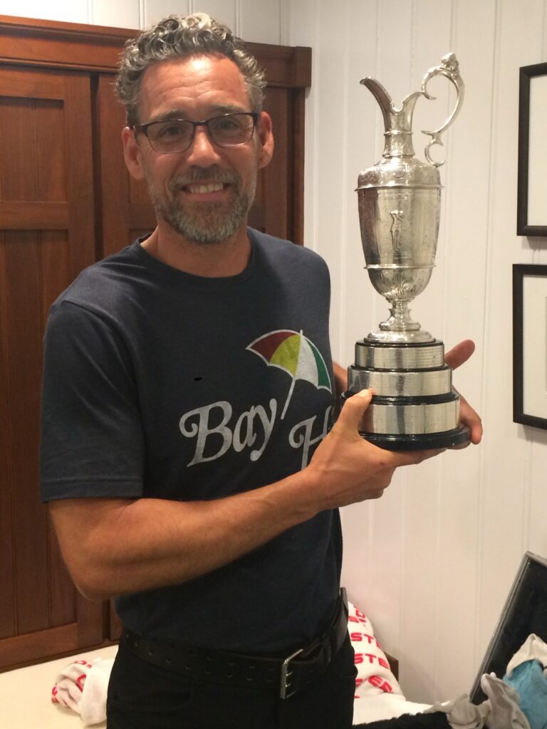 Man in a "Bay' H" shirt holds the Claret Jug trophy.