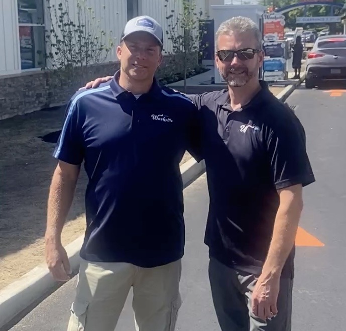 Two men in uniform stand together outside a car wash, one wearing a hat and the other sunglasses.