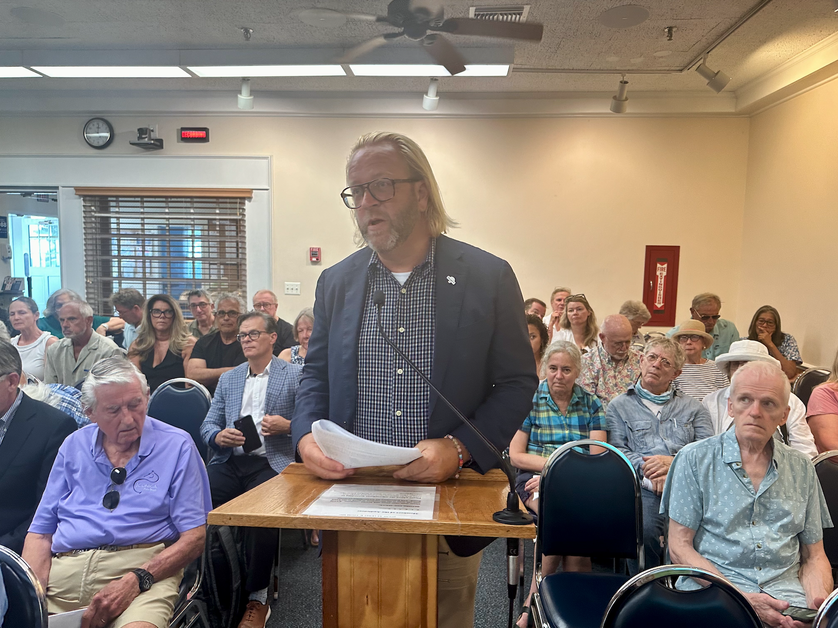 A man with long blonde hair and thick-rimmed glasses stands speaking at a podium at a public hearing.