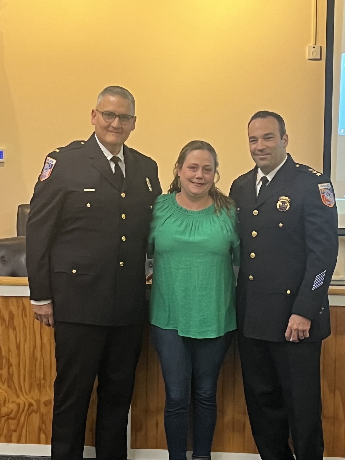 A woman in a green shirt stands between two uniformed policemen, posing for a photo together to celebrate a police promotion.