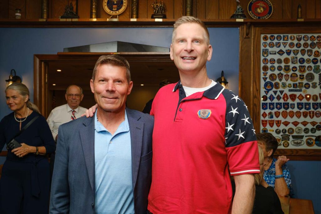 Two white men stand arm in arm posing and smiling for the camera
