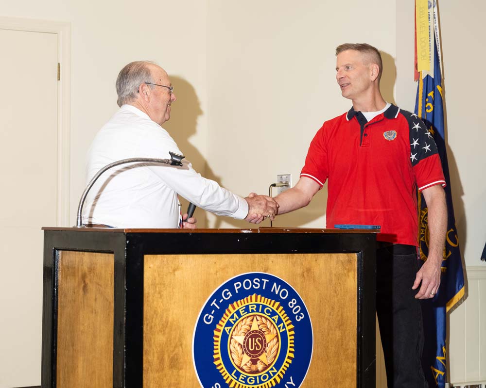 Two white men shake hands behind a podium. One holds a microphone.