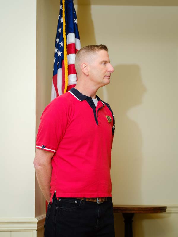 A white man in a red shirt stands with his hands behind his back, in front of an American flag.