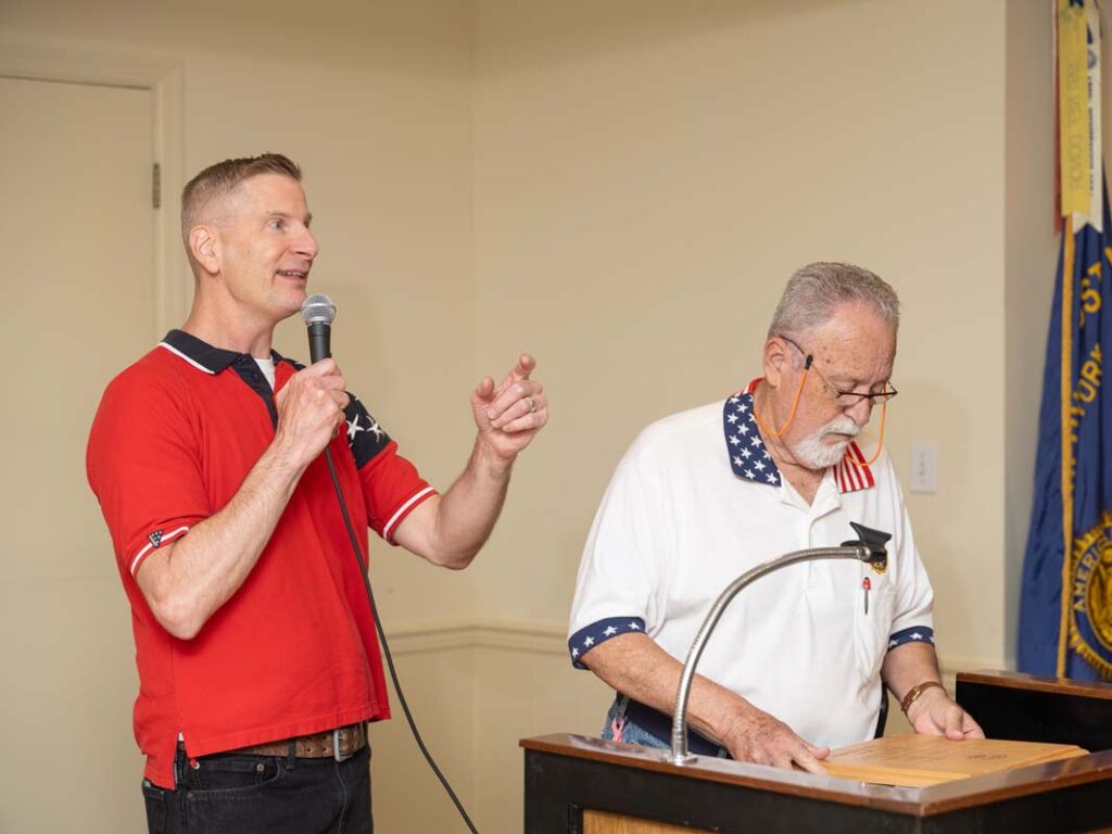 Two white men stand at a podium. One is speaking into a microphone; the other is looking down at the wooden plaque he is about to present to the speaker.