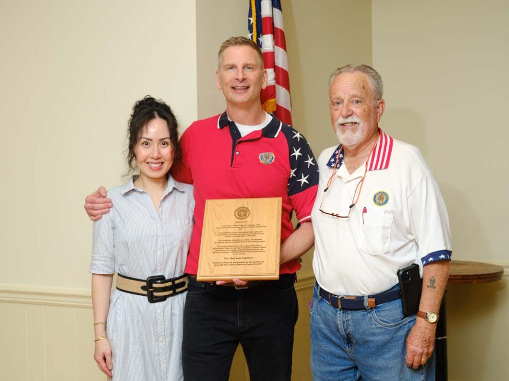 One white woman and two white men stand in a line. The man in the middle holds a wooden plaque honoring his service to the town of Southold.