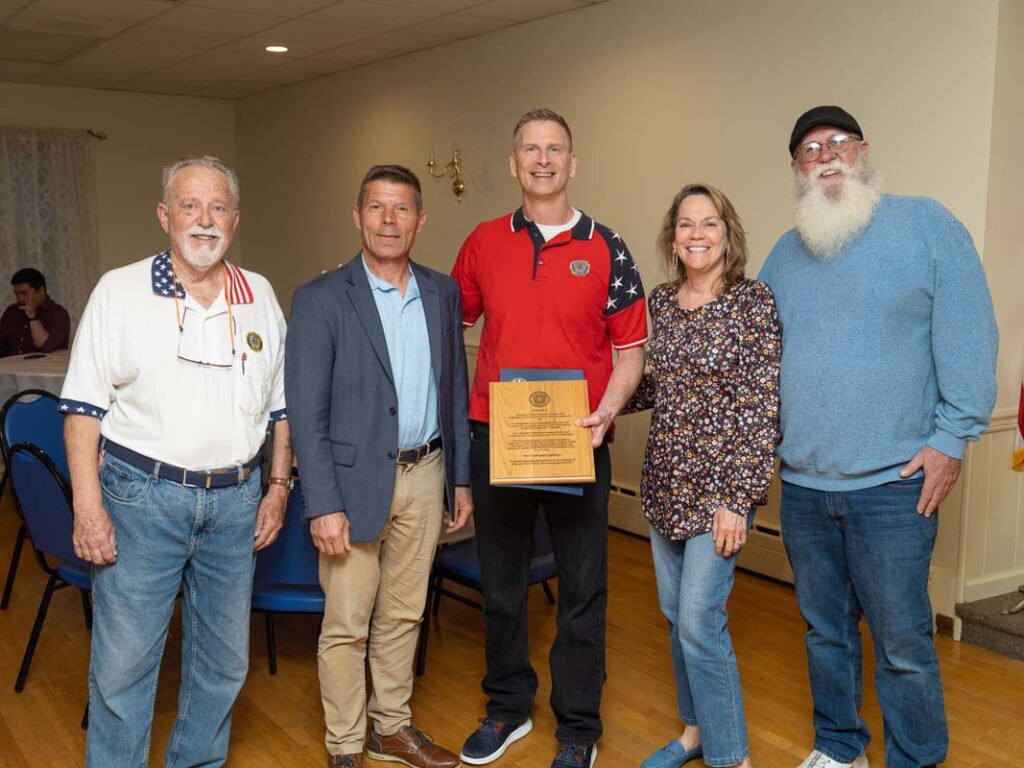 Five white people stand in a line. The one in the middle hold a plaque and a blue envelop holding a proclamation