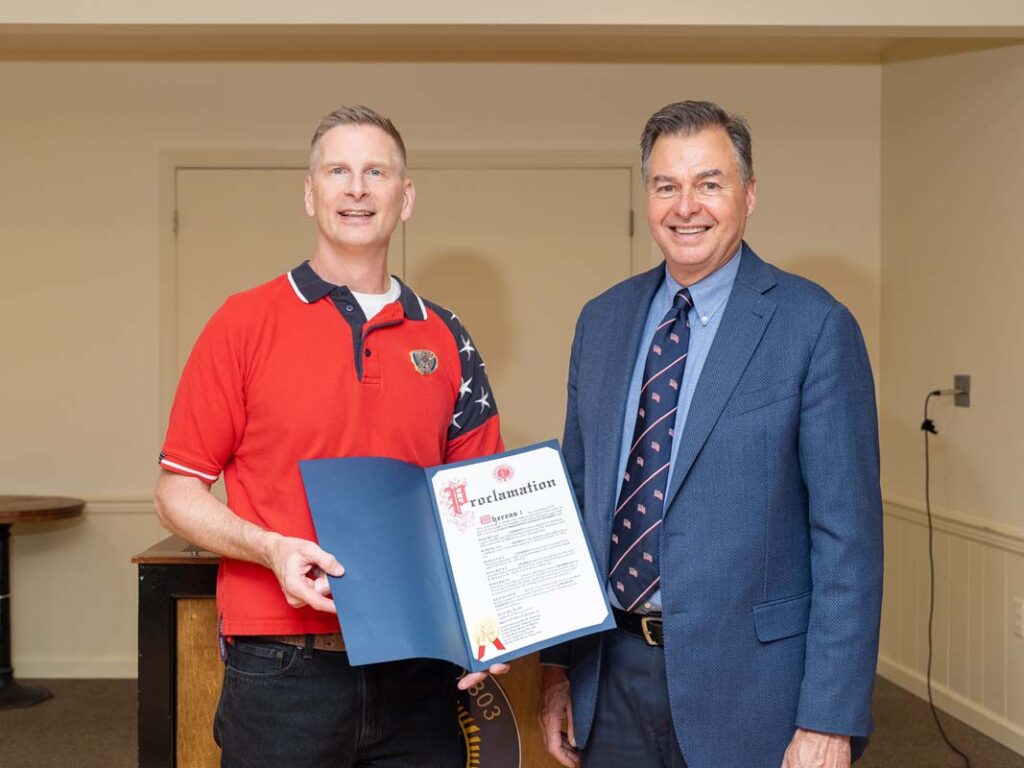 Two white men stand side by side. One holds a blue envelop open showing the proclamation he received from the town of Southold