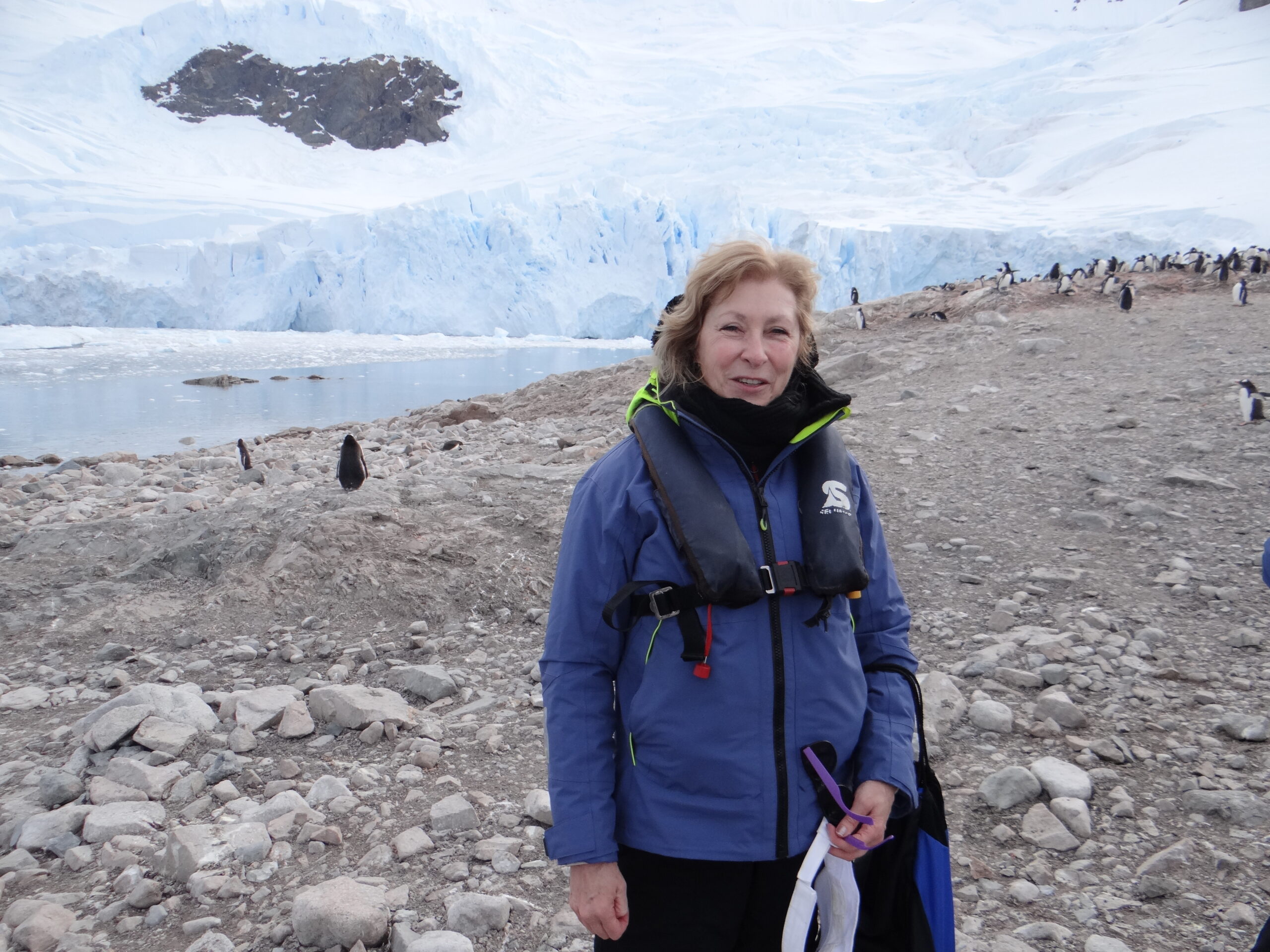 A woman stand dressed in a big puffy parka, and ventures forth in Antarctica with penguins in the background.