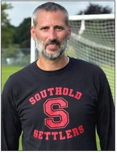 A man, Chris Ruggi, has a salt-and-pepper beard and hair, and wears a black, long-sleeved shirt with a red Southold Settlers logo on it.