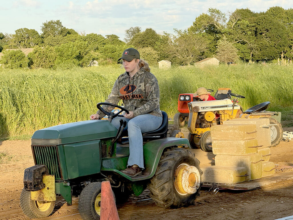 A girl in a camouflage hooded sweatshirt, jeans and baseball cap pulls the weighted sled with her green tractor.