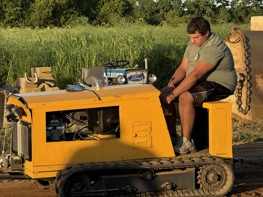 A boy in a green shirt drives a yellow tractor with track-type wheels, not tires.