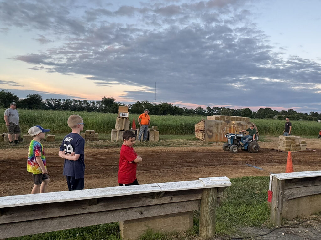 Three boys stand on the tractor pull track watching as the winner drives his blue tractor in a victory lap.