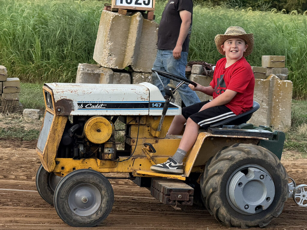 A boy in a red T-shirt, black shorts and a straw hat sits on a yellow tractor.