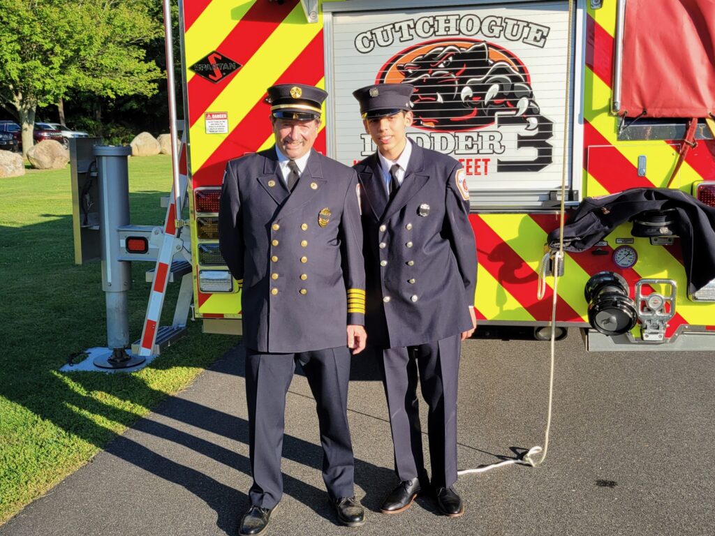A grandfather firefighter and grandson firefighter pose in their dress uniforms in front of the back of a fire engine.