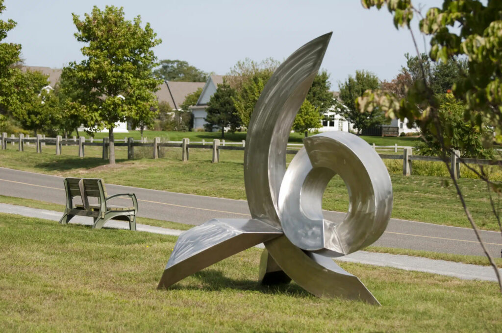A modern metal sculpture stands on a grassy lawn next to a park bench and a paved path.