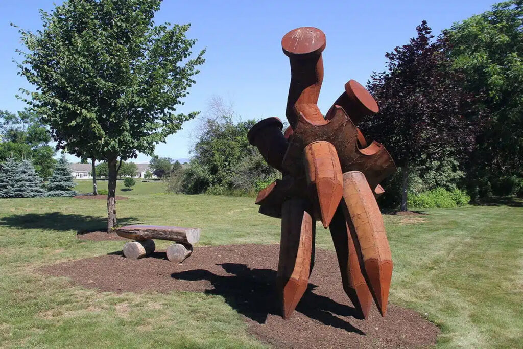 A rustic metal sculpture stands in a park next to a log bench under a tree.