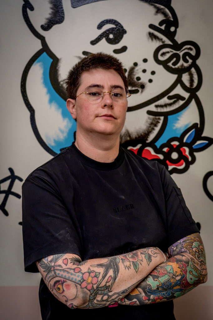 A person with short hair and glasses, wearing a black t-shirt with "SLICER" on it, poses with their arms crossed in front of a mural.