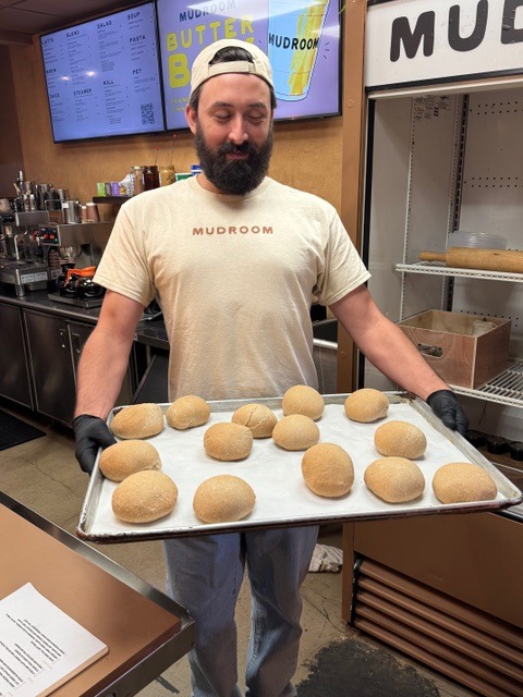 Man in a "MUDROOM" shirt holds a baking sheet with 12 bread rolls.